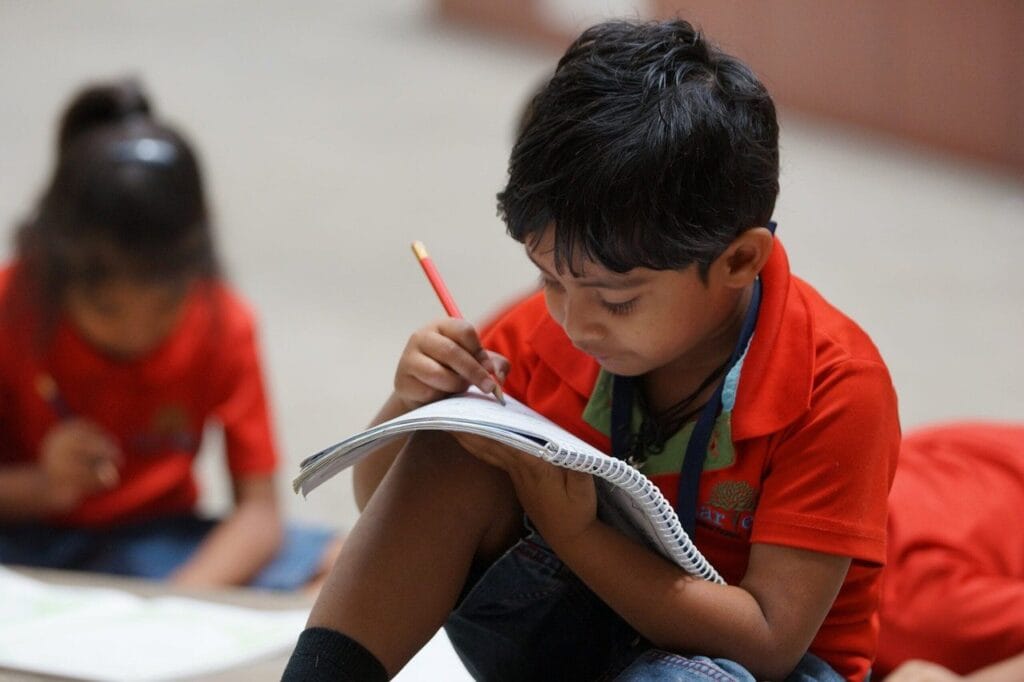 Child writing in notebook with pencil, another child in background.